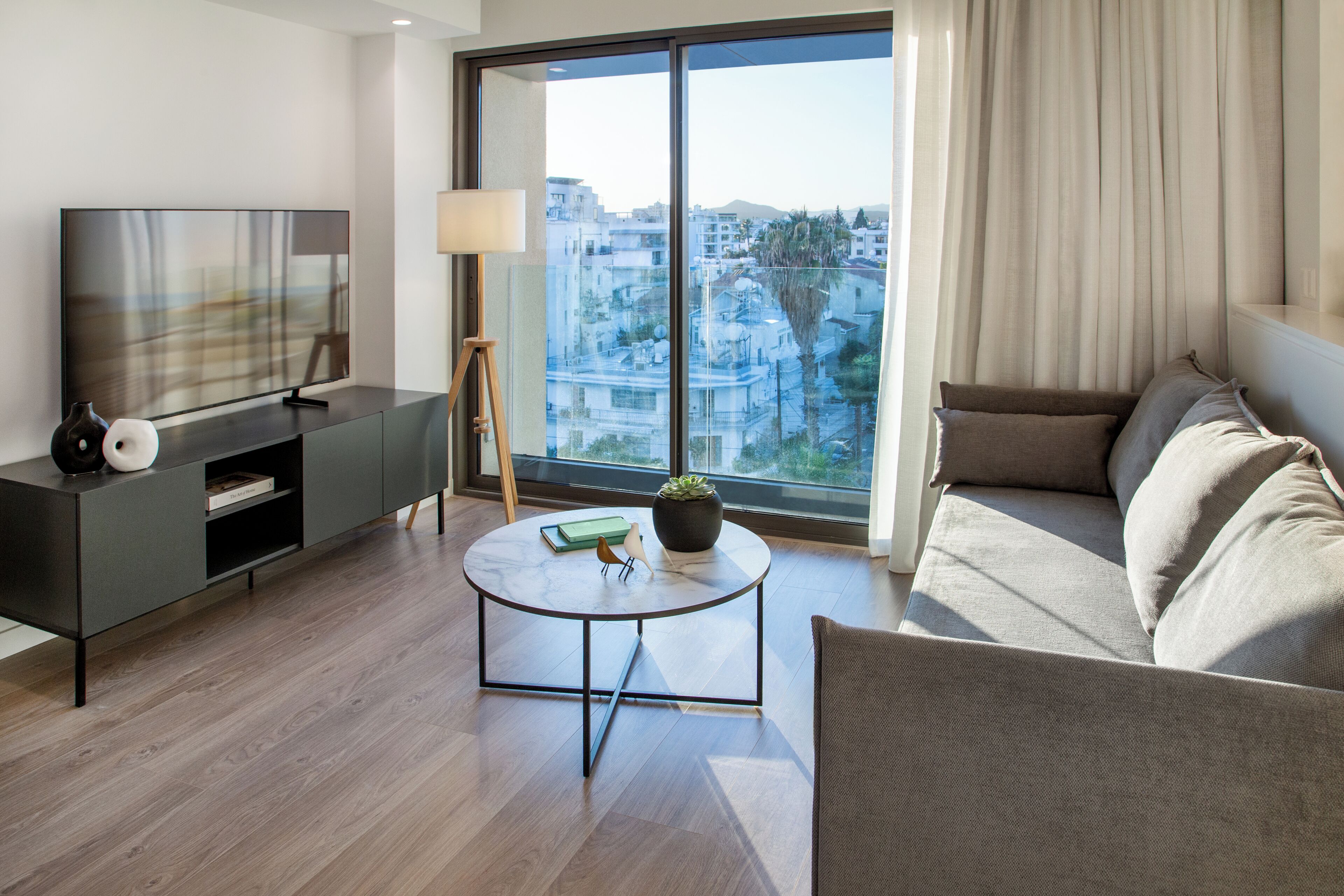A modern living room with a TV, sofa, and a large window offering a city view.