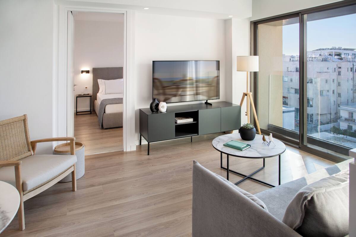Una habitación de hotel moderna con un ventanal grande, un televisor de pantalla plana, un sofá, una mesita y una cama con una mesita de noche.