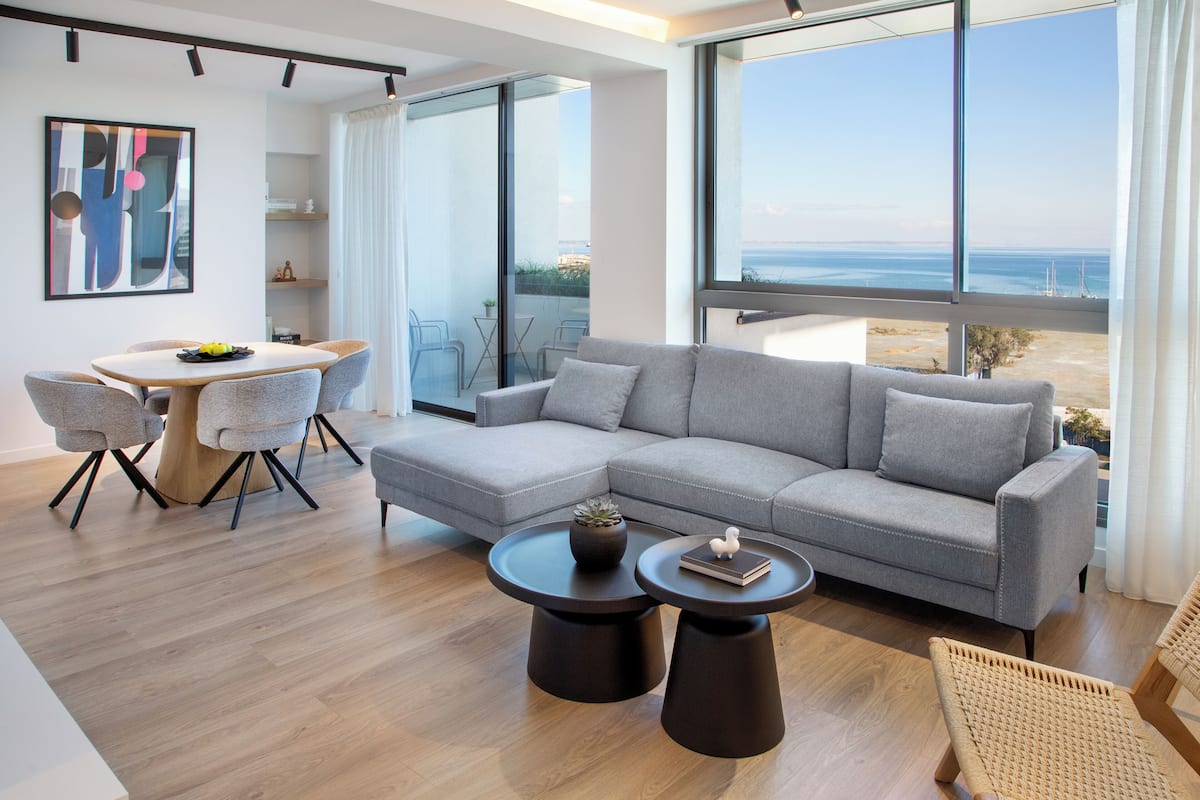 Una sala de estar moderna con un sofá seccional gris, dos mesas redondas de centro, una mesa de comedor con sillas y un ventanal con vistas al mar.