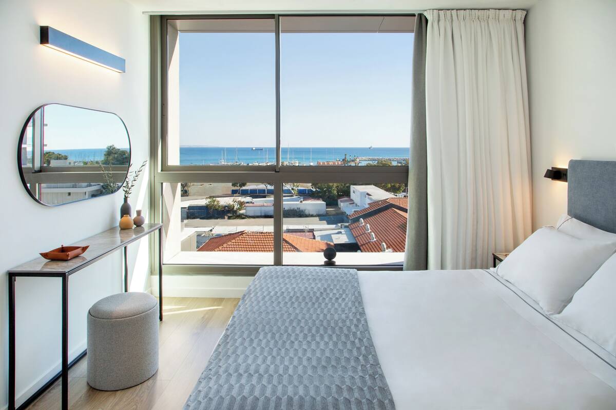 Habitación de hotel con un ventanal grande, una cama, una mesita de noche, un espejo y vistas al mar y edificios.