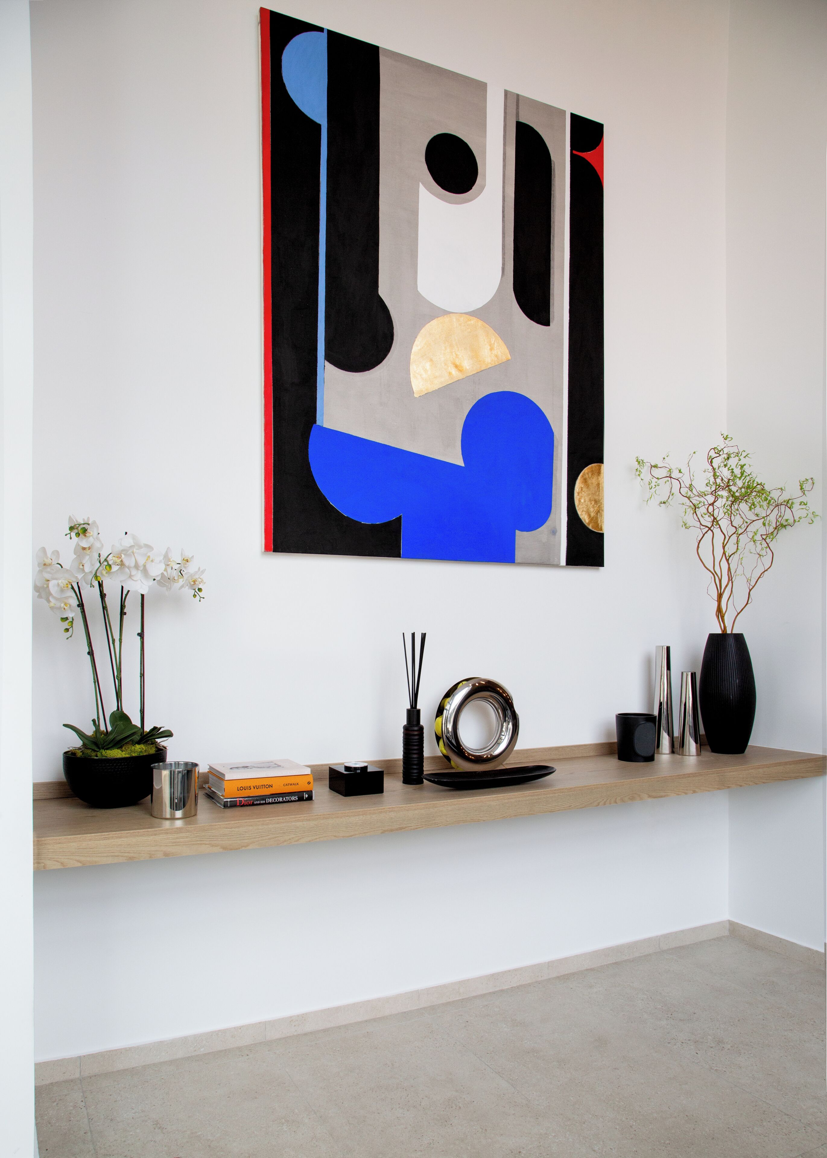 A modern interior with a minimalist art piece, a potted orchid, a vase with branches, and a shelf with decorative items.