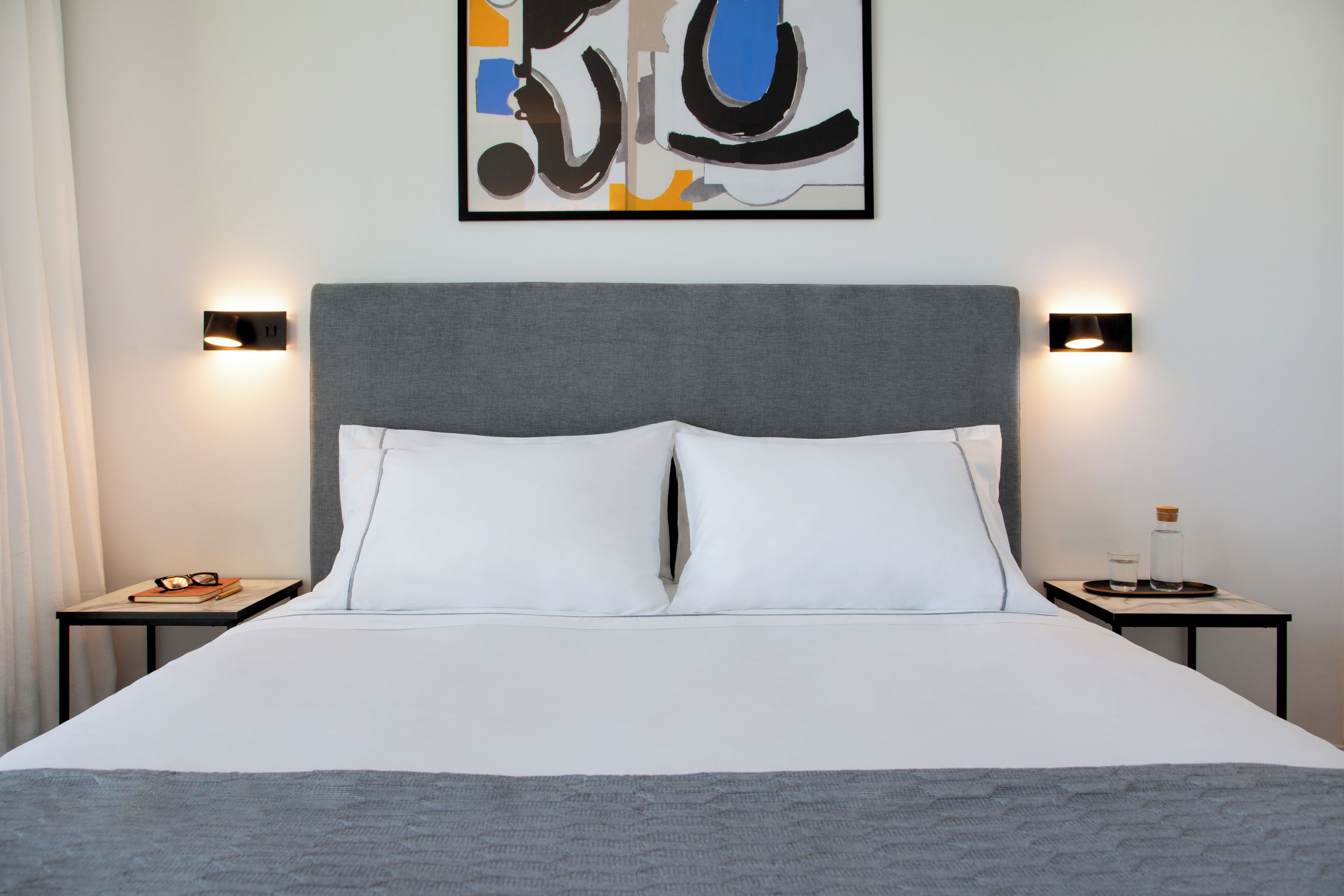Un lit bien fait, avec du linge de lit blanc, deux tables de chevet avec des lampes et une œuvre d’art abstraite encadrée accrochée au mur.