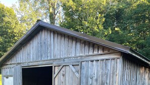 A wooden barn with a large open door, surrounded by grass and trees.
