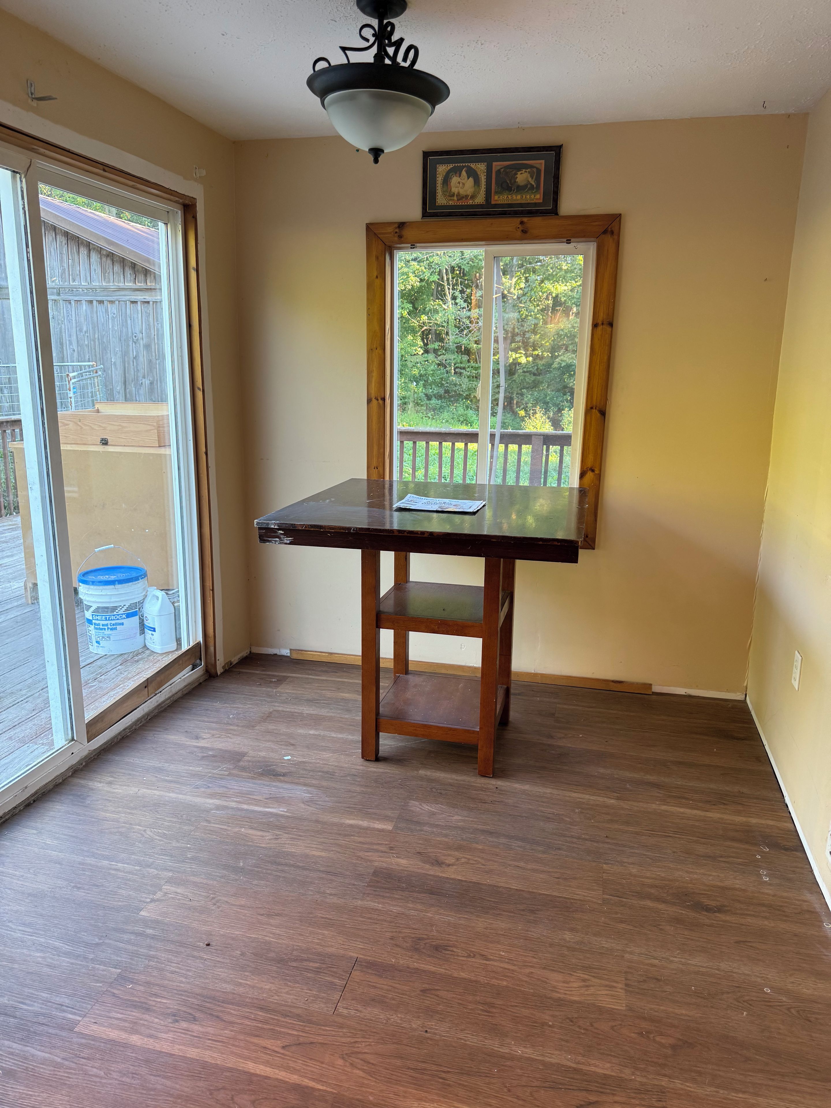 A room with a wooden table, a ceiling light fixture, and a window with a view of a wooden deck and greenery.
