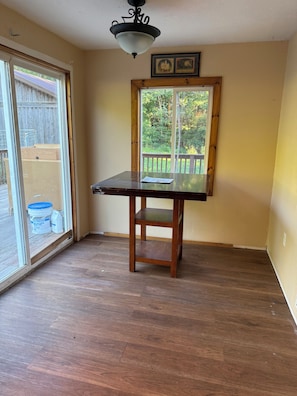 A room with a wooden table, a ceiling light fixture, and a window with a view of a wooden deck and greenery.
