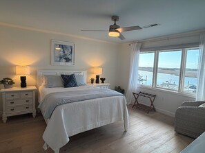 Ein Schlafzimmer mit Bett, Nachttischen, Deckenventilator, Fenster mit Blick auf das Wasser und einem kleinen Tisch am Fenster.