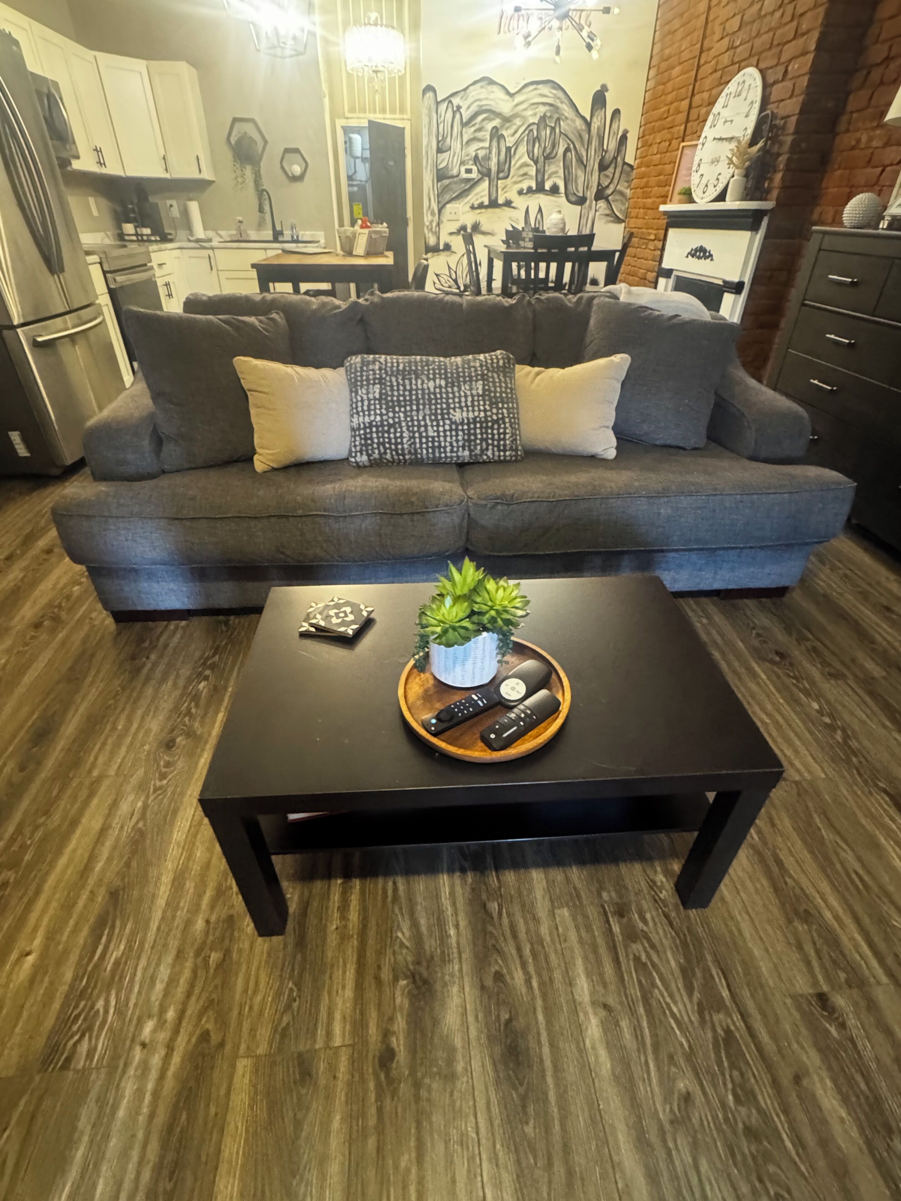 A living room with a dark brown coffee table, a grey sofa with patterned cushions, a small potted plant, and a remote control tray.