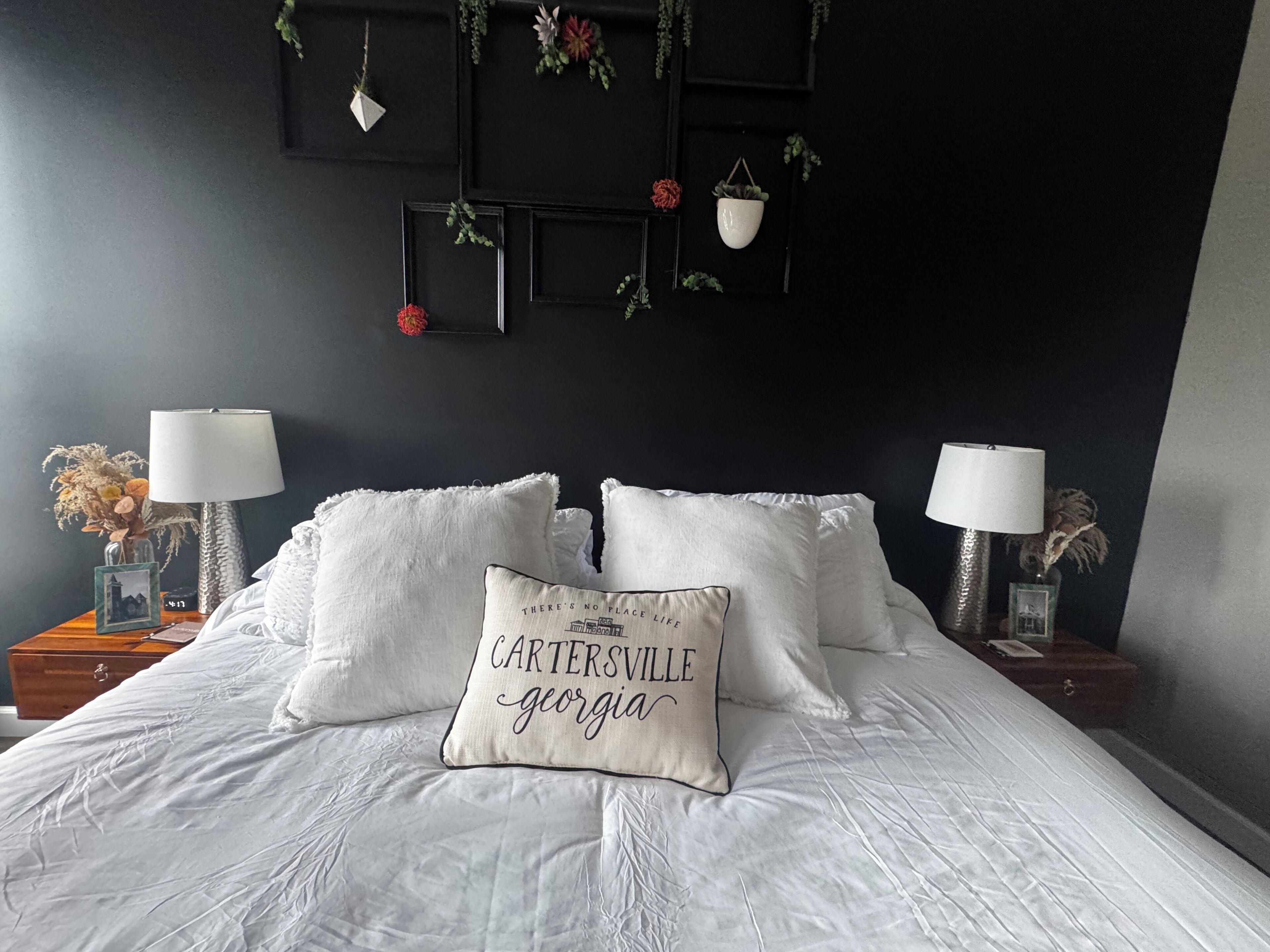 A bedroom with a bed, two bedside tables with lamps, a wall-mounted shelf with decorative items, and a pillow with text.