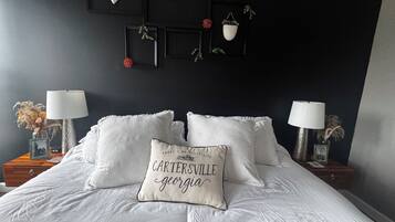 A bedroom with a bed, two bedside tables with lamps, a wall-mounted shelf with decorative items, and a pillow with text.