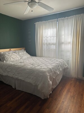 A bedroom with a bed, wooden flooring, a ceiling fan, and a window with curtains.