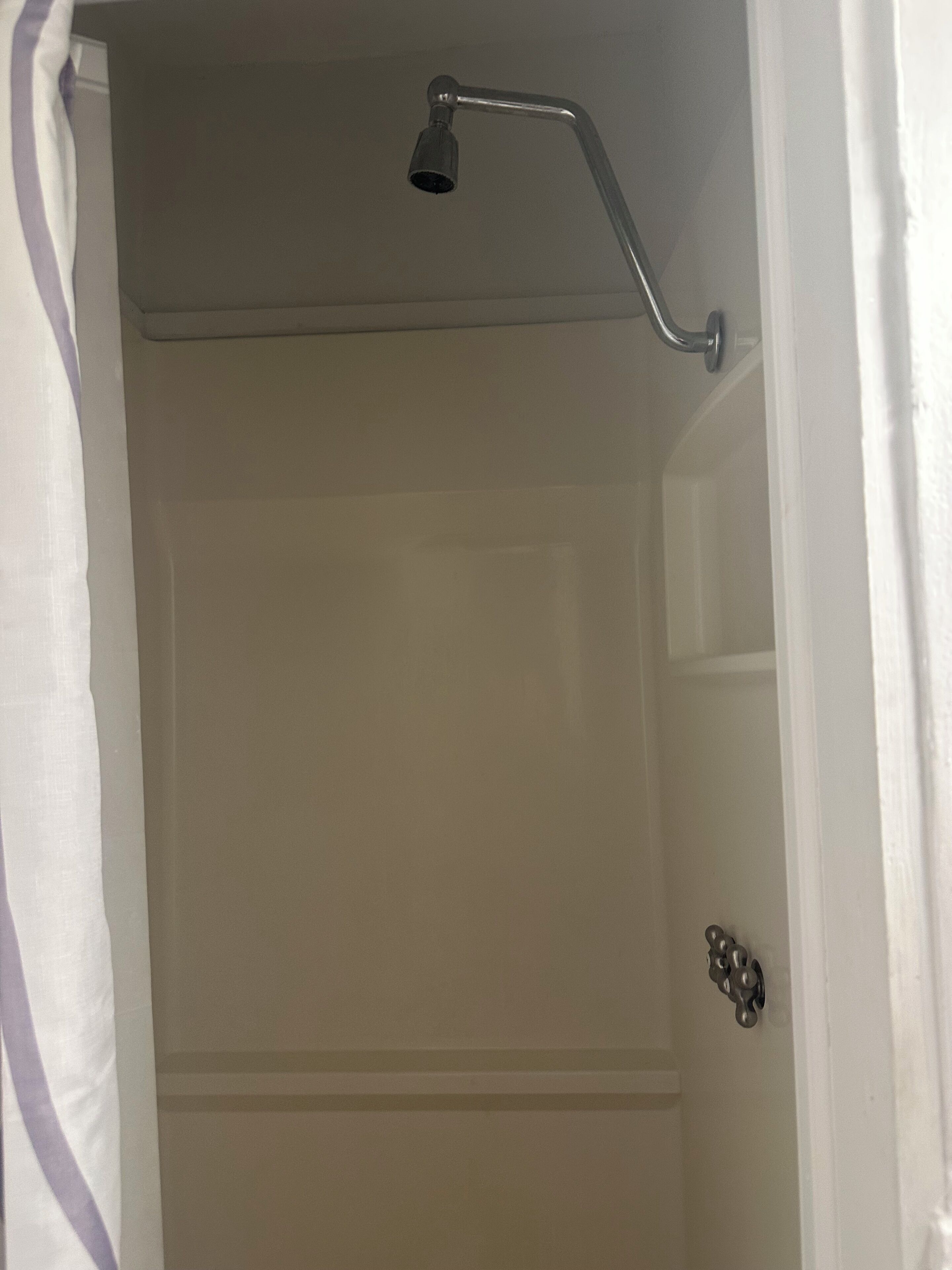 Une douche avec un pommeau de douche amovible, une porte avec une poignée ronde et un rideau blanc.