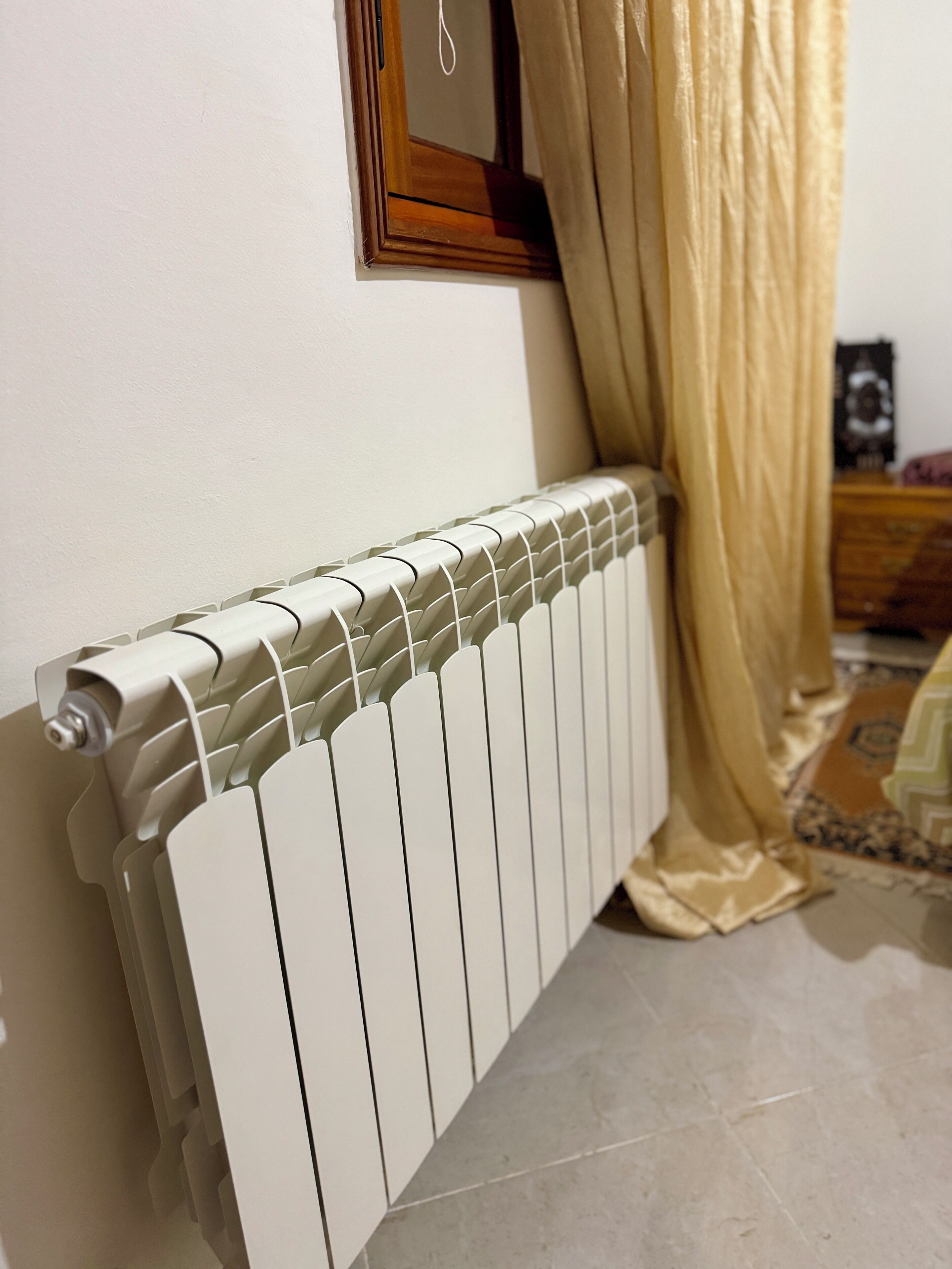 Un radiateur blanc dans une pièce dont le sol est carrelé, avec une commode en bois en arrière-plan.