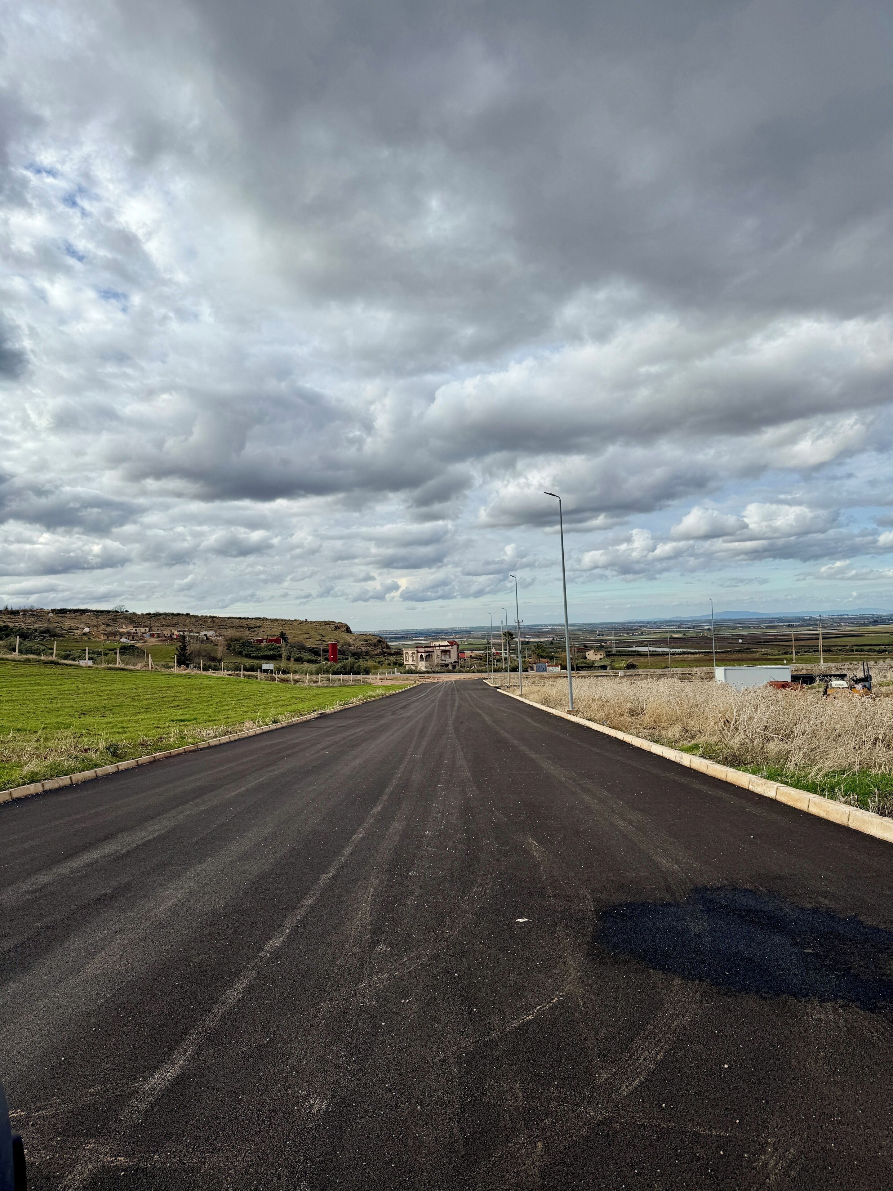 A rural road with a clear sky, distant buildings, and open fields.