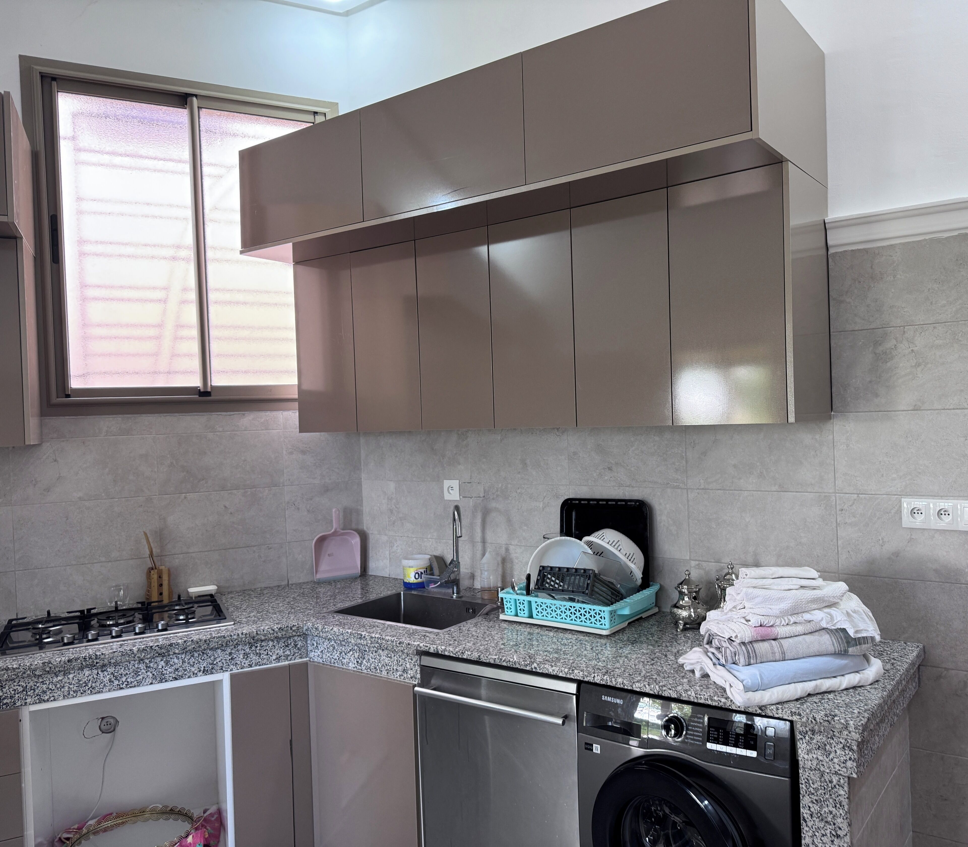 Una cocina con estufa a gas, lavabo, escurreplatos, lavadora y ventana con persianas.