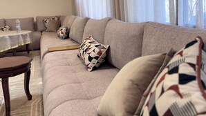 A living room with a sofa, pillows, a coffee table, and a view of the outdoors through sheer curtains. - Maison de charme en ferme privée- Atlas (El Hajeb)