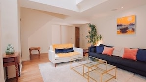 Un salon avec un canapé blanc, un canapé noir, une table basse en verre, un tapis blanc, une table d’appoint en bois et un tableau accroché au mur.