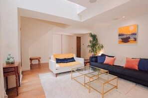 A living room with a white sofa, a black sofa, a glass coffee table, a white rug, a wooden side table, and a painting on the wall. - Niru Large Double Living Room Maisonette (London)