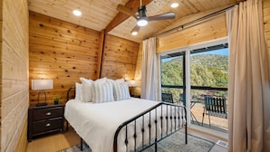 Een slaapkamer in een houten chalet met een bed, nachtkastje, lamp en uitzicht op een balkon met buitenmeubilair.