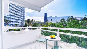A balcony with a white railing, a small round table with a plate of fruit, two chairs, and a view of a cityscape with buildings and greenery. - Sea View Studio Playa de Las Americas (Playa de Las Americas)