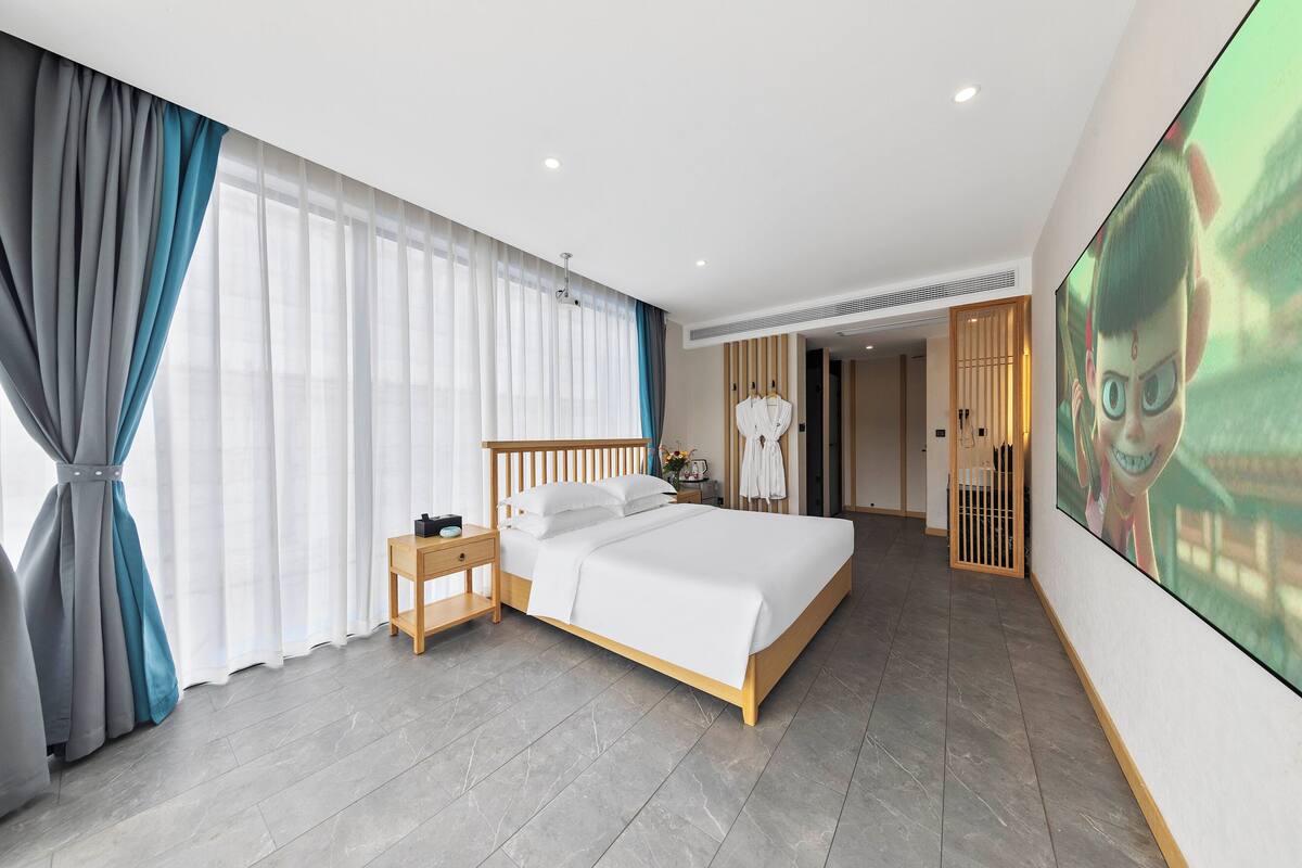 Una habitación de hotel moderna con una cama grande, una mesita de noche, un cabecero de madera, un ventanal con cortinas traslúcidas, un televisor montado en la pared y una puerta de madera que da a un baño.