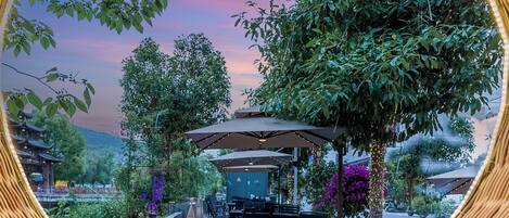 An outdoor seating area with umbrellas, tables, and chairs, surrounded by greenery and a view of a traditional building.