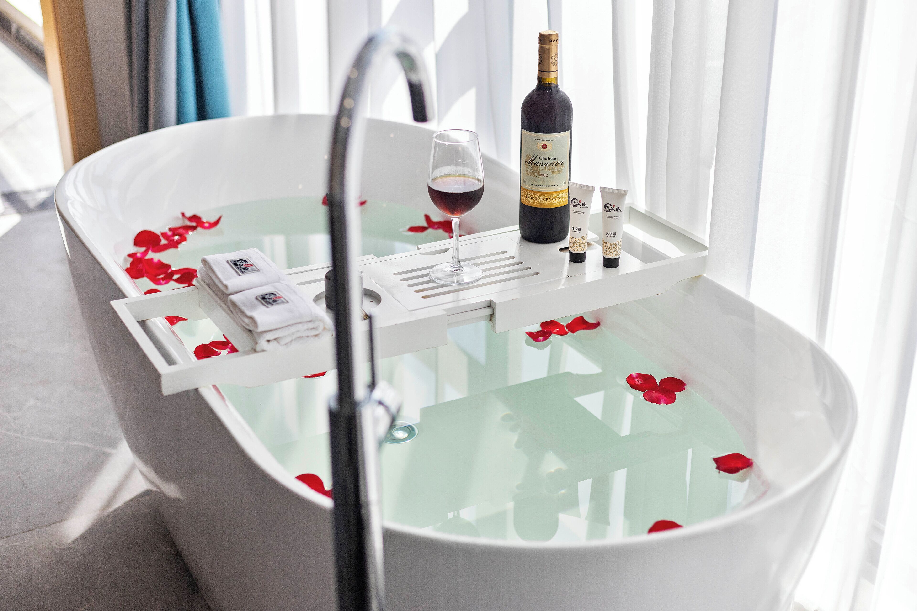 A bathtub with rose petals, a bottle of wine, and toiletries.