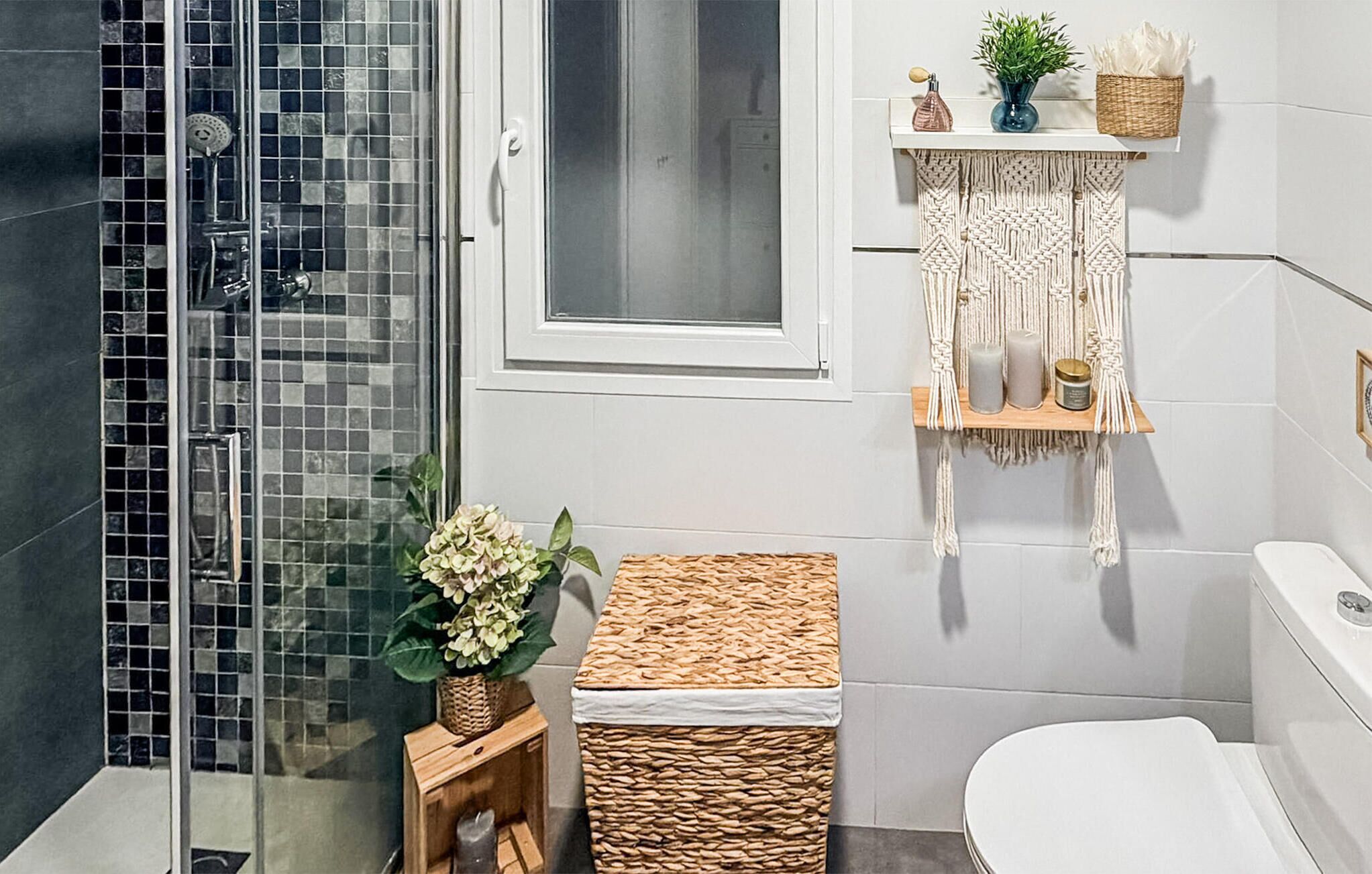 A bathroom with a glass shower enclosure, a white toilet, a woven basket, and a macrame shelf with candles.