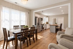 Cozinha moderna com eletrodomésticos de aço inoxidável, sala de jantar com cadeiras de madeira e sala de estar com sofá estampado.