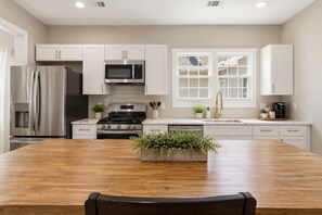 A modern kitchen with stainless steel appliances, white cabinetry, a wooden dining table, and a window with blinds. - The Runway Retreat - 8 minutes to ATL Airport! (Atlanta)