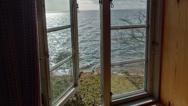Ein Zimmer mit zwei geöffneten Fenstern mit Blick auf ein Gewässer, einem Holzboden und einem kleinen Holzgegenstand auf dem Boden.