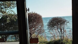 Blick durch ein Fenster auf eine Wiese mit Tisch, Stühlen, Bäumen und einem Gewässer.