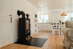 Een kamer met een open haard, houten vloer, witte wanden, een eethoek met stoelen en een hanglamp.
