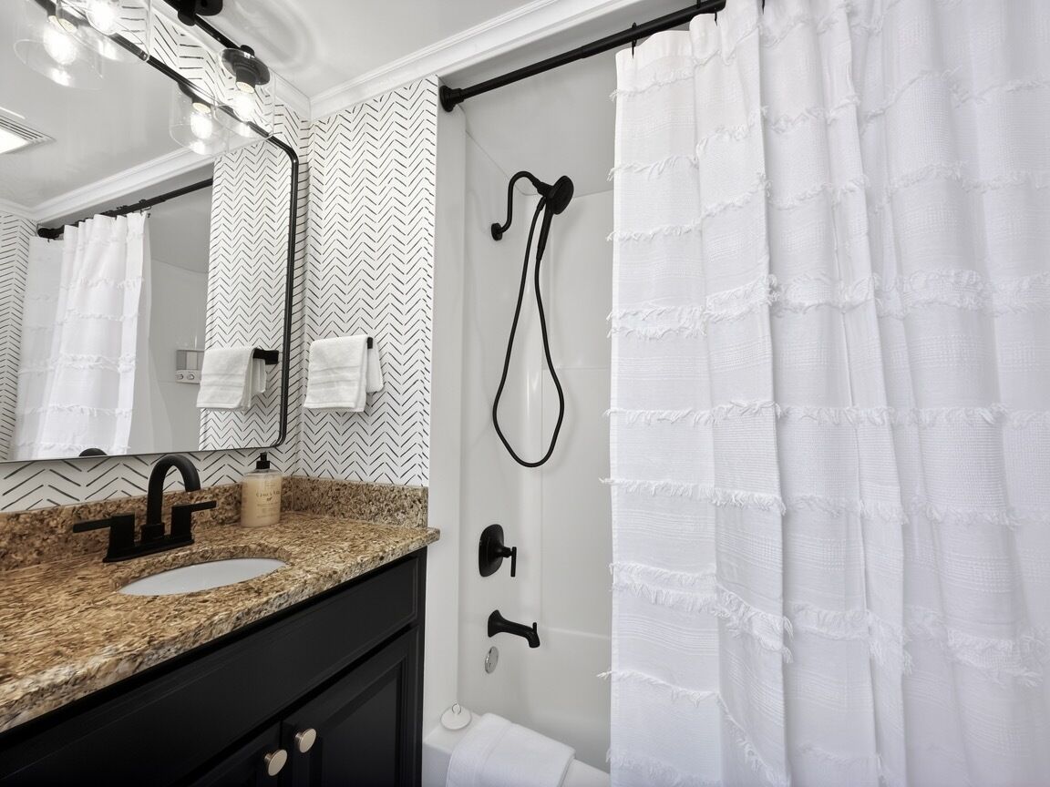 A bathroom with a granite countertop, a black faucet, a white sink, a mirror, a shower with a white curtain, and a towel rack.