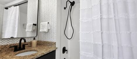 A bathroom with a granite countertop, a black faucet, a white sink, a mirror, a shower with a white curtain, and a towel rack.