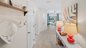 A hallway with a white door, a hat rack with a hat, a shelf with a 'happy place' sign, a lamp, and a painting on the wall. - Bright Beach Condo, Pool, Spa, Comfy Beds, Patio (St. Augustine)