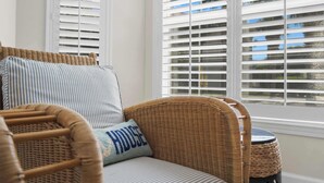 A wicker chair with a cushion labeled 'HOUSE' and a striped pillow, positioned near a window with white blinds. - Bright Beach Condo, Pool, Spa, Comfy Beds, Patio (St. Augustine)