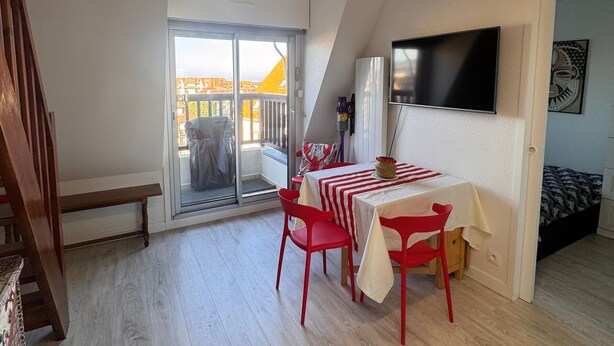A small, open-plan living space with a balcony, a dining table with red chairs, a bed, and a flat-screen TV mounted on the wall. - Bright duplex near the beach - 4 guests (Villers-sur-Mer)