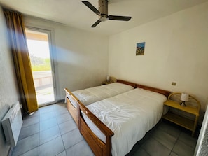 Un dormitorio con cama, ventilador de techo, una mesita de noche con lámpara y ventana con cortinas.