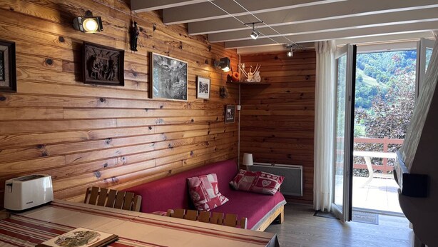 Ein gemütliches Zimmer mit Holzwänden, einem roten Sofa, einem Esstisch mit kariertem Tischläufer und Blick ins Grüne.