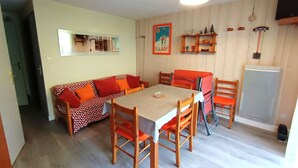 Et spiseområde med et bord og stole, en sofa, et køkkenområde med hylder og et køleskab.