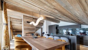 Eine moderne Küche mit Holzbalkendecke, einem Essbereich mit langem Tisch und Stühlen sowie einer sichtbaren Kochinsel mit Geräten.