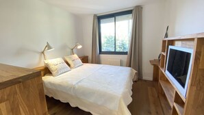 Un dormitorio con una cama de madera, dos lámparas de noche, un ventanal con cortinas y un televisor sobre un mueble de madera.