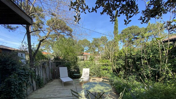 Una terraza de madera que da a un jardín con mesa y sillas, rodeado de árboles y una cerca.
