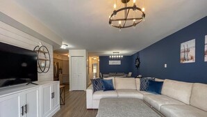 A living room with a sectional sofa, a flat-screen TV, a chandelier, and a kitchen in the background. - Harbor Front Condo. Minutes to downtown. (Cape May)
