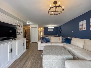 A living room with a sectional sofa, a flat-screen TV, a chandelier, and a kitchen in the background. - Harbor Front Condo. Minutes to downtown. (Cape May)
