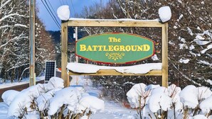Un letrero de madera con la inscripción 'The Battlefield', rodeado de ramas cubiertas de nieve.