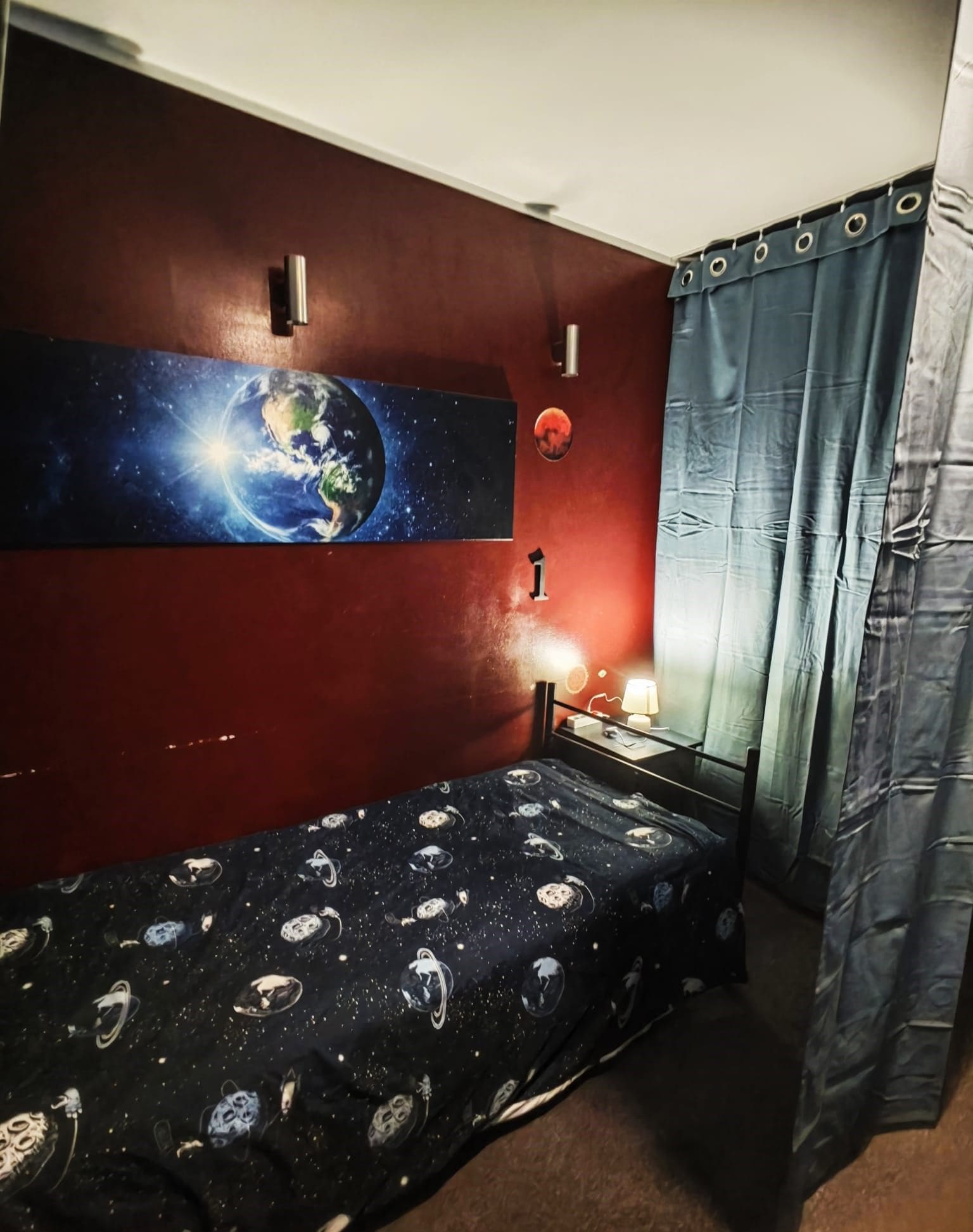 Une pièce avec un lit, un téléviseur fixé au mur, une lampe et un rideau.