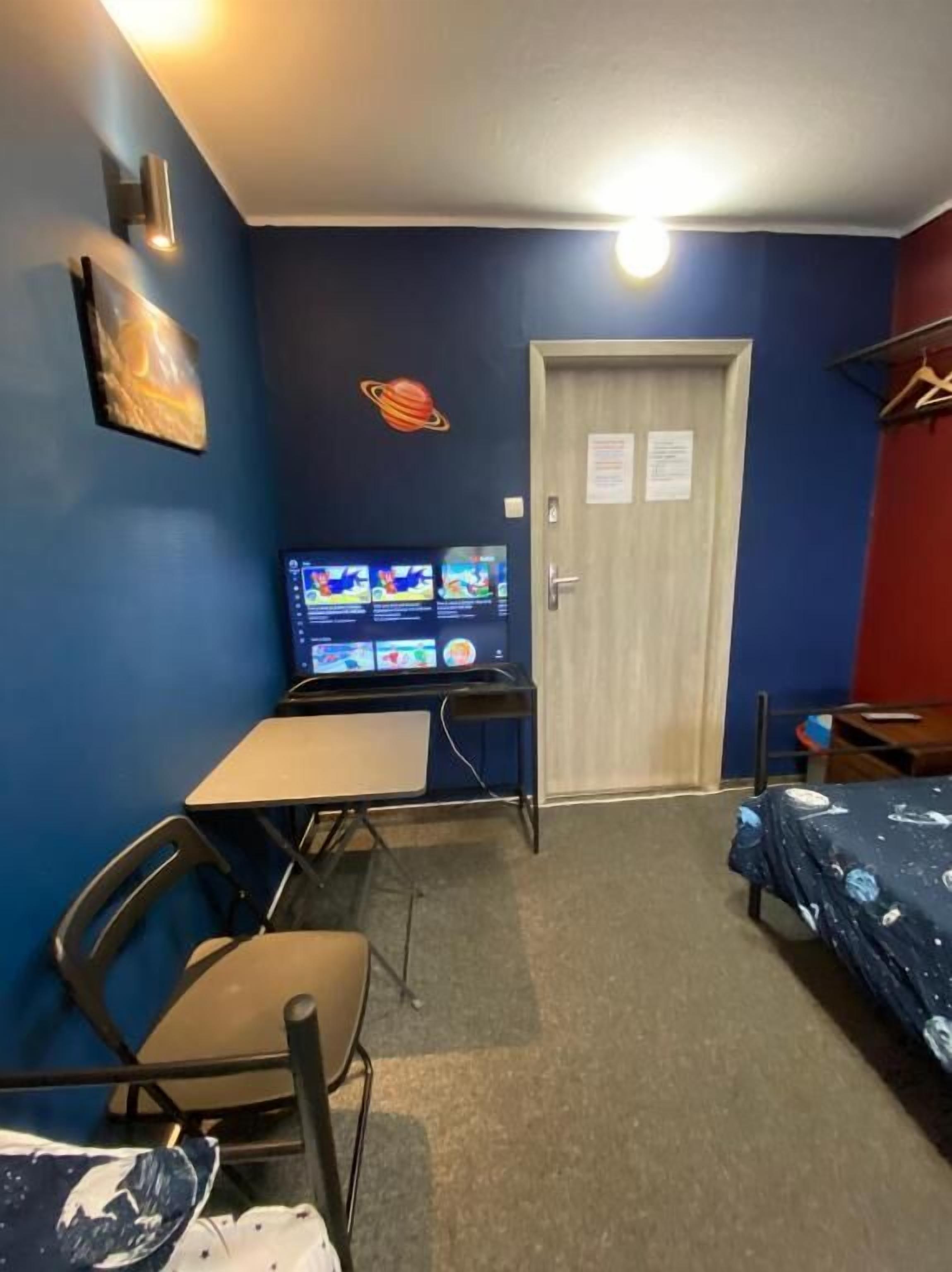 Quarto com escrivaninha, duas cadeiras, cama com colcha de tema espacial, TV na parede e porta com aviso.