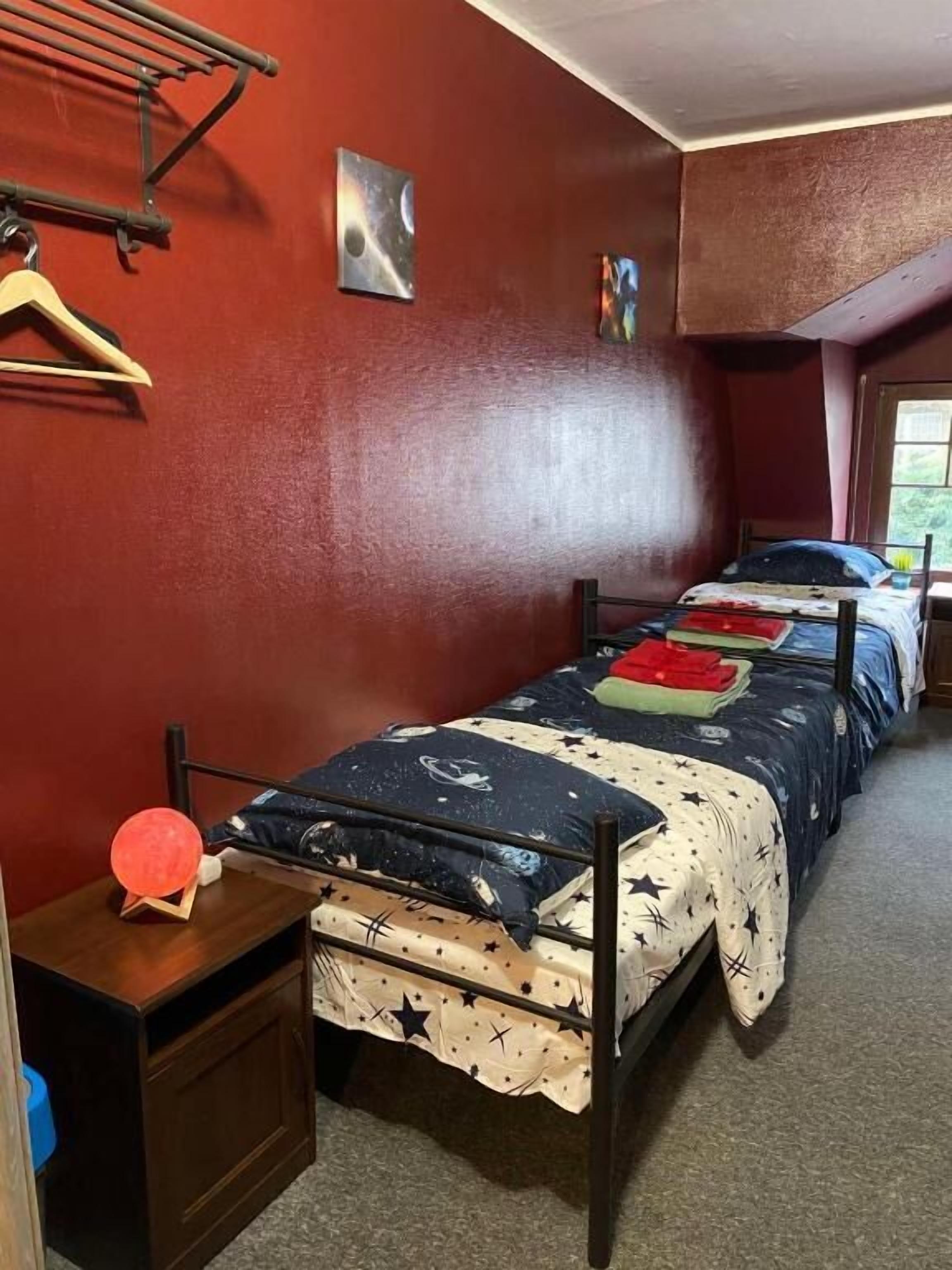 Un dormitorio con dos camas individuales, una mesita de noche de madera, una lámpara roja y un tendedero de pared.