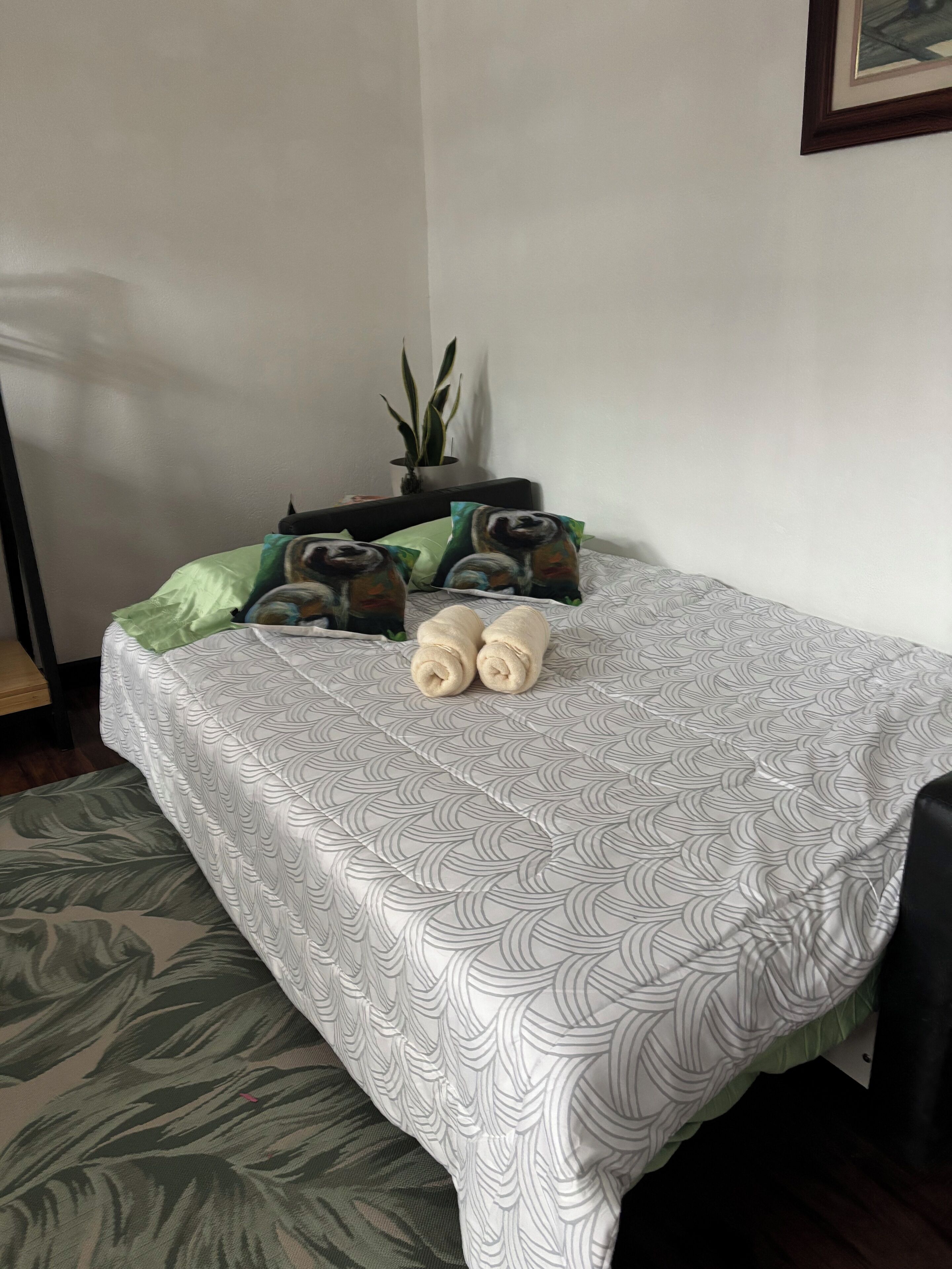 Un lit bien fait, avec une couvre-lit à motifs, deux serviettes enroulées et deux oreillers décorés d’un motif de paresseux.